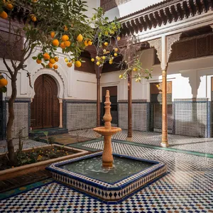 A traditional Moroccan riad courtyard with detailed zellige tilework, flowing fountain, citrus trees, and ornate woodwork. Morning light casting intricate shadow patterns. - Ideogram Ideogram V2