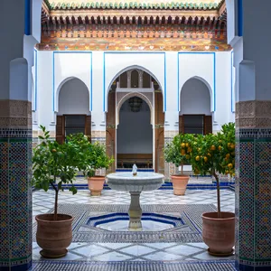 A traditional Moroccan riad courtyard with detailed zellige tilework, flowing fountain, citrus trees, and ornate woodwork. Morning light casting intricate shadow patterns. - Reve Reve Image (Halfmoon)