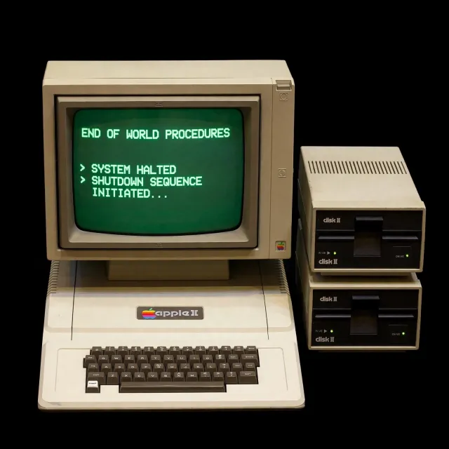 Vintage Apple II computer with green monochrome CRT screen, displaying 'END OF WORLD PROCEDURES' in green text. Two external floppy drives stacked on the right, labeled disk II with rainbow Apple logos. Beige casing, black background, retro aesthetic. - Muse Image