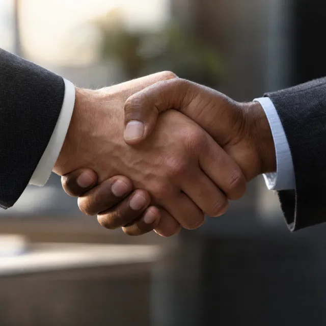 A close-up of two people shaking hands, both hands fully visible with five fingers each, in sharp focus against a blurred background. - Takumi 1.1