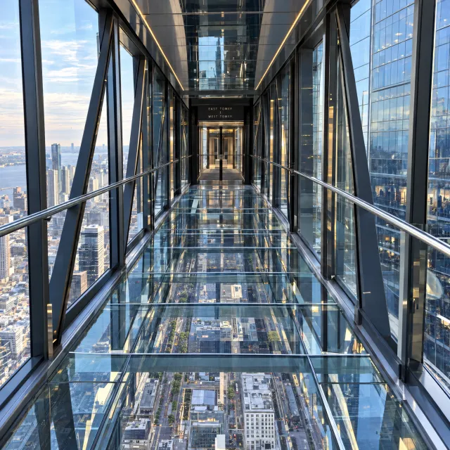 Photorealistic rendering of a contemporary glass skybridge between two skyscrapers, seen from inside with city views visible through glass walls and floor. Show accurate reflections and structural details. - Takumi 1.1