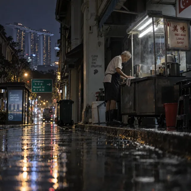 Rain-slicked Singapore street, 3 AM. A lone elderly hawker cleans his cart under one flickering fluorescent light. Steam rises gently. Low-angle shot, photorealistic - Takumi 1.1