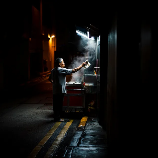 Rain-slicked Singapore street, 3 AM. A lone elderly hawker cleans his cart under one… - Reve Image (Halfmoon)